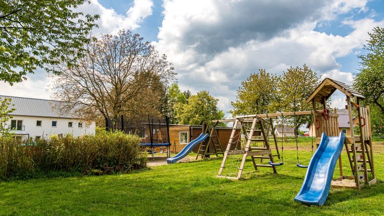 Ein Spielplatz mit einer Rutsche und Schaukeln in einem grünen Garten. Im Hintergrund sind Bäume und ein Himmel mit einigen Wolken sichtbar.
