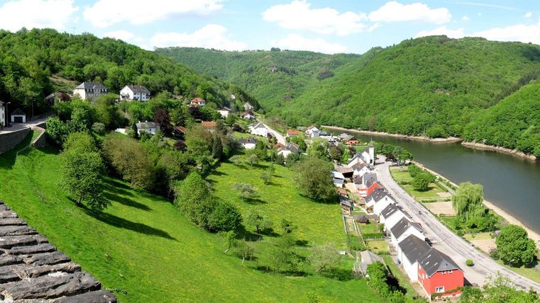 Une vallée pittoresque avec une rivière, entourée d'un greenery luxuriant et de petites maisons. Le ciel est clair et bleu, créant une atmosphère paisible.