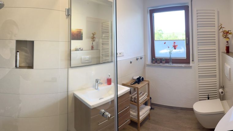 Une salle de bain moderne avec une douche et une grande fenêtre. Les murs sont clairs, et il y a des éléments décoratifs comme des fleurs et des images.