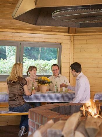 Een groep van vier personen zit in een gezellige hut en geniet van de tijd samen. Op de tafel staat een boeket bloemen en op de achtergrond brandt een open haard.