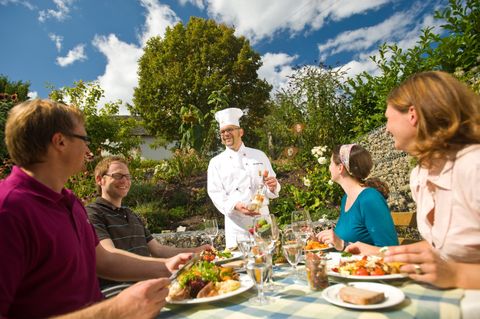Ein Koch serviert ein köstliches Essen im Freien. Eine Gruppe von Menschen genießt das Essen in einer schönen Gartenumgebung.