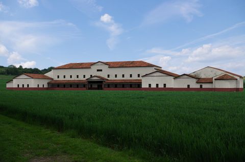 Ein großes, modernes Gebäude mit einem roten Dach vor einem grünen Feld. Der Himmel ist blau mit einigen Wolken.
