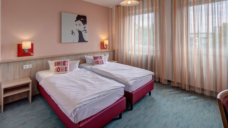 Une chambre moderne avec deux lits simples et une palette de couleurs chaudes. De grandes fenêtres avec des rideaux laissent entrer beaucoup de lumière naturelle.