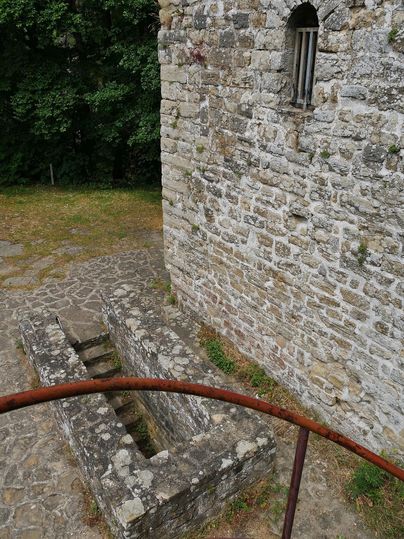 Ein historisches Mauerwerk mit Steinen und einem kleinen Fenster. Im Vordergrund sind alte Steinstrukturen auf dem Boden sichtbar.