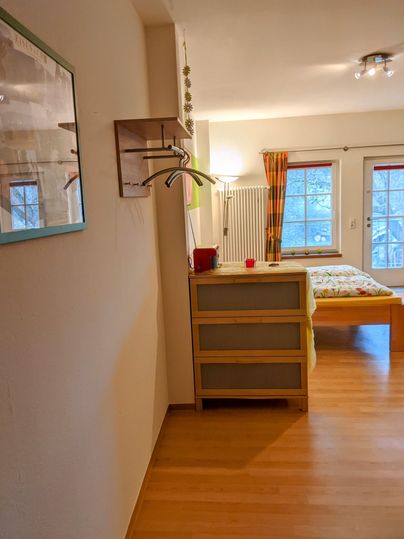 Ein gemütliches Zimmer mit Holzfußboden und einem Bett im Hintergrund. An der Wand befinden sich ein Spiegel und ein Kleiderhaken.