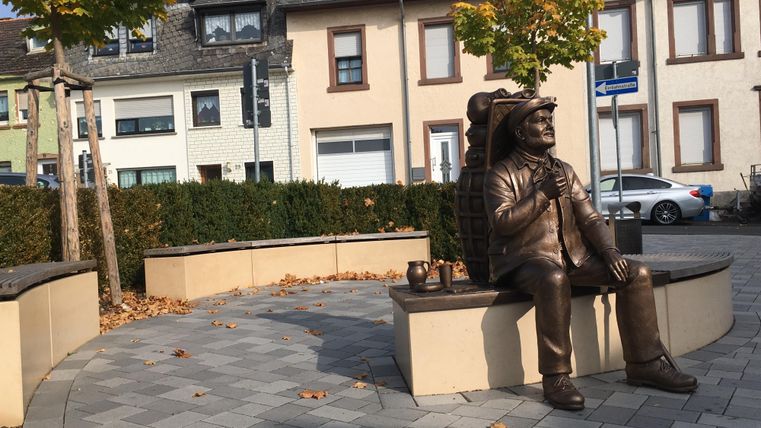Eine Bronzestatue eines Mannes sitzt auf einer Bank. Um ihn herum sind Bäume und moderne Gebäude zu sehen.
