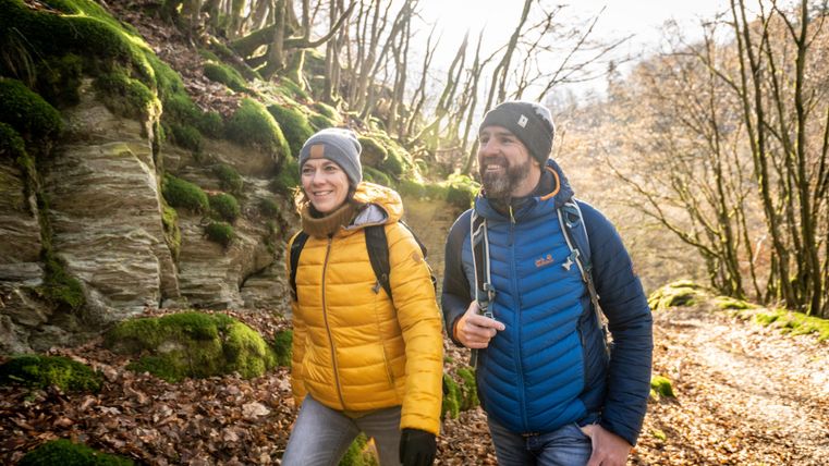 Zwei Wanderer auf einem schönen Waldweg mit grünem Moos und Felsen. Es scheint ein sonniger Tag zu sein und die Natur um sie herum ist lebendig.