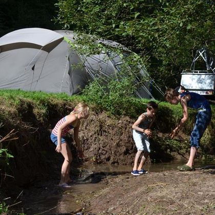 A tent stands by the edge of a small stream where children are playing. The sun is shining and the surroundings are green and natural.