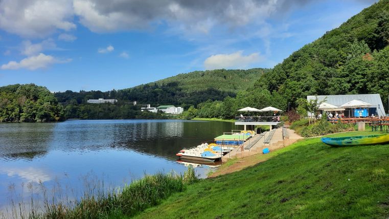 Ein ruhiger See umgeben von grünen Hügeln. In der Nähe gibt es eine Terrasse mit Tretbooten und Sitzmöglichkeiten.