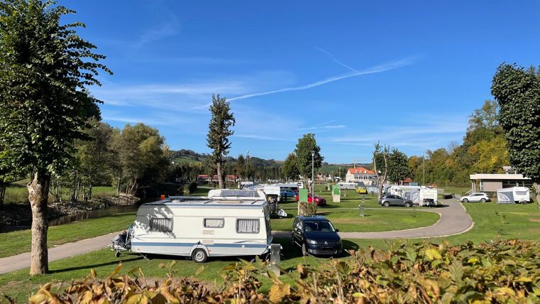 Ein Campingplatz mit mehreren Wohnwagen und Autos in einer grünen Umgebung. Der Himmel ist blau und die Sonne scheint.
