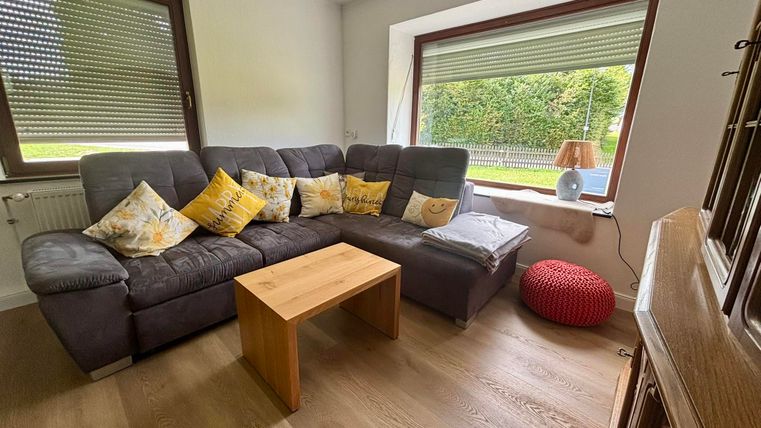 Ein gemütliches Wohnzimmer mit einem grauen Sofa und bunten Kissen. Ein Holztisch steht in der Mitte und ein Fenster bietet Blick auf den Garten.