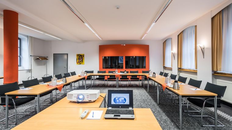 Een moderne vergaderruimte met tafels en stoelen in U-vorm. Aan de wand bevindt zich een projector en een scherm, en de ruimte heeft grote ramen met daglicht.