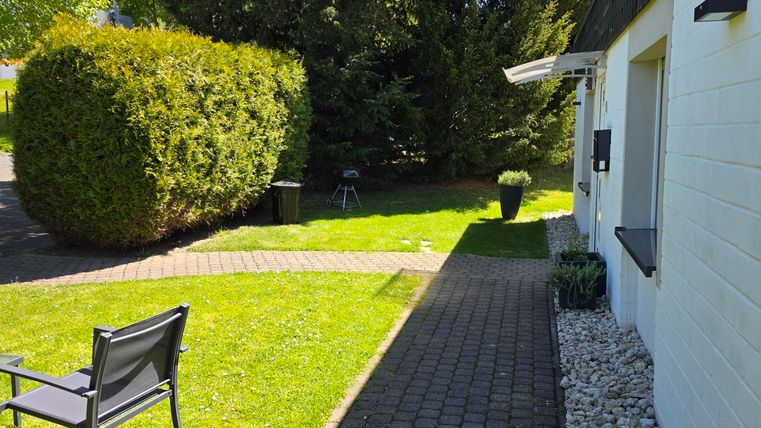 Ein gepflegter Garten mit grünem Rasen und Büsche. Auf dem Weg steht ein Gartenstuhl und die Sonne scheint.