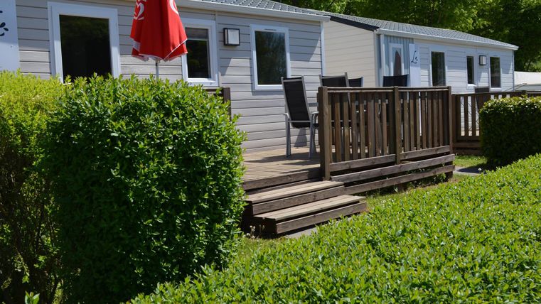 Ein modernes Mobilheim mit Holzveranda und Gartenmöbeln. Im Vordergrund befinden sich gepflegte grüne Büsche.