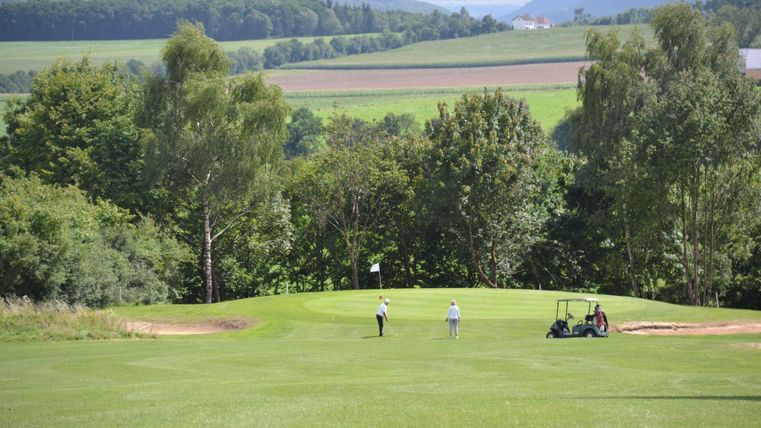 Eine weitläufige Golfanlage mit grünen Fairways und Bäumen im Hintergrund. Zwei Golfer sind auf dem Platz, während ein Golfwagen in der Nähe steht.