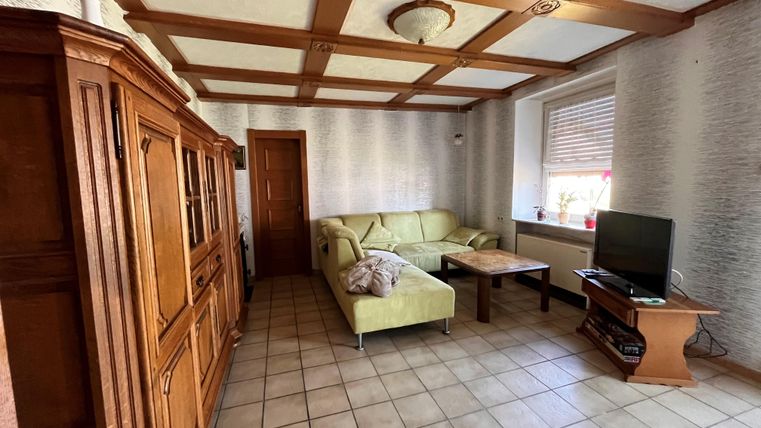 Ein gemütliches Wohnzimmer mit einem hellgrünen Sofa und einem Holztisch. An der Wand steht ein Fernseher und ein Holzschrank ist sichtbar.