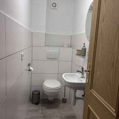 Een moderne badkamer met een witte betegelde wand en een toilet. Er is een handwasbak en een spiegel.