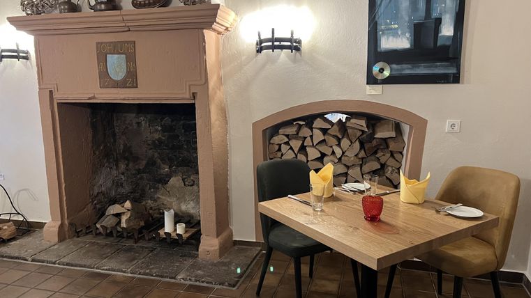 Un espace restaurant confortable avec une cheminée et un tas de bois. Sur la table se trouvent des serviettes jaunes et un verre rouge.