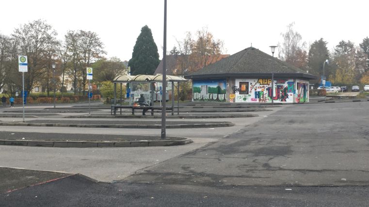Een lege parkeerplaats met een kleine, kleurrijke gevel op de achtergrond. Het is bewolkt en enkele bomen zijn in de buurt zichtbaar.