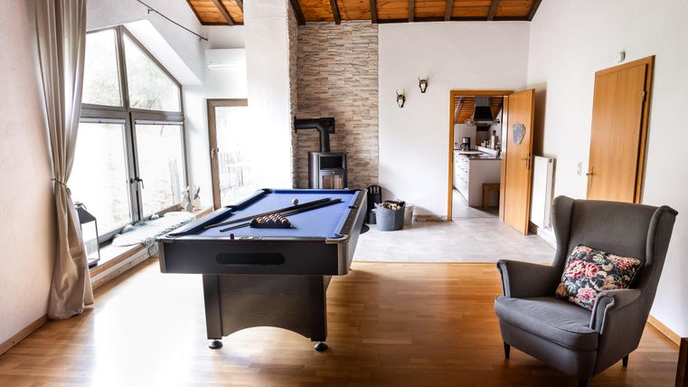 A bright room with a pool table in the middle and large windows. A cozy armchair is located against the wall.