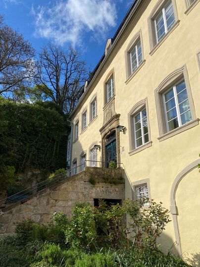 Ein historisches Gebäude mit gelber Fassade und großen Fenstern. Im Vordergrund sind grüne Pflanzen und ein paar Stufen sichtbar.