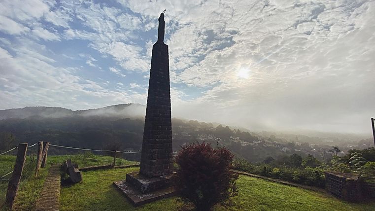 Ein obeliskartiges Denkmal steht auf einer grünen Wiese mit Blick auf das Tal. Sanfte Hügel und bewölkter Himmel umgeben die Szene.