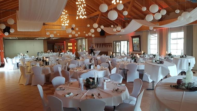 Een feestelijk versierde ruimte met ronde tafels en een witte tafelkleed. Het plafond is versierd met heldere bollen en stoffen banen.