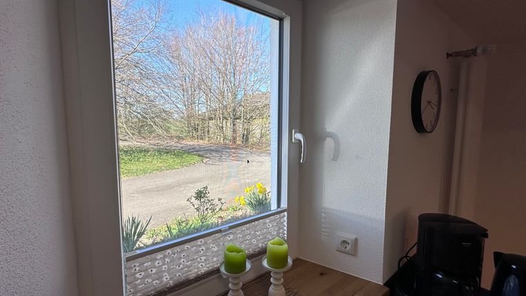 Ein helles Fenster mit Blick auf einen Garten und einen klaren blauen Himmel. Auf der Fensterbank stehen zwei Kerzen und eine Uhr an der Wand.
