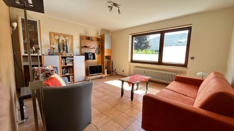 Ein geräumiges Wohnzimmer mit einer orangenen Couch und einem grauen Sessel. Große Fenster lassen viel Licht herein und zeigen einen Blick ins Freie.