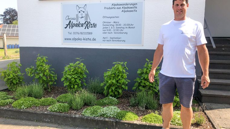 Un homme se tient devant un bâtiment à la façade blanche. Il porte un t-shirt et un short, et autour de lui se trouvent des plantes vertes.
