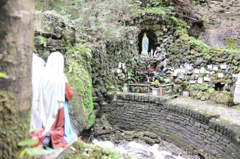 Eine kleine Kapelle in der Natur mit einer Statue von Maria. Grüne Pflanzen und ein klarer Bach umgeben den Ort.
