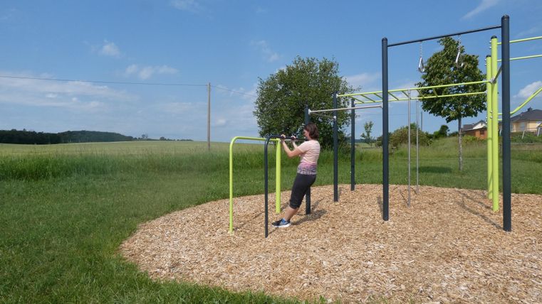 A person is training on a fitness machine outdoors. In the background, green meadows and a clear blue sky can be seen.