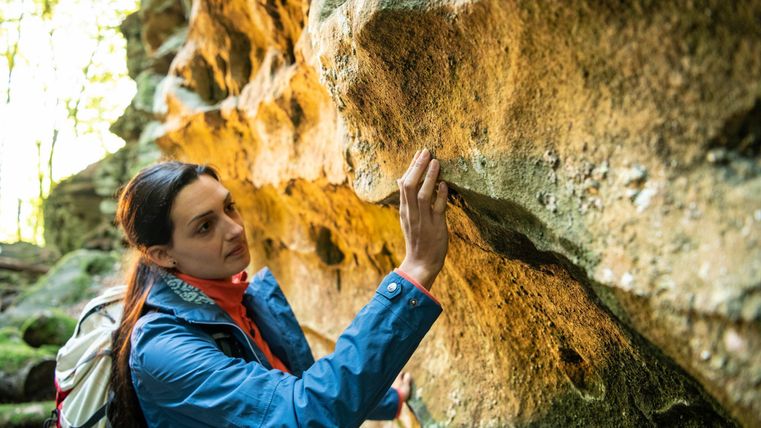 Eine Frau untersucht eine Felswand in einem Waldgebiet. Sie trägt eine Jacke und einen Rucksack und scheint interessiert an der Natur um sie herum zu sein.