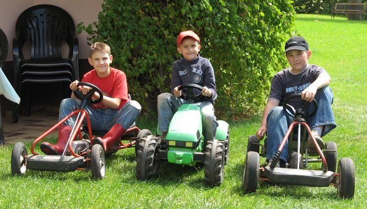 Trois enfants sont assis sur des Kettcars dans une prairie verte. Ils sourient et s'amusent ensemble.
