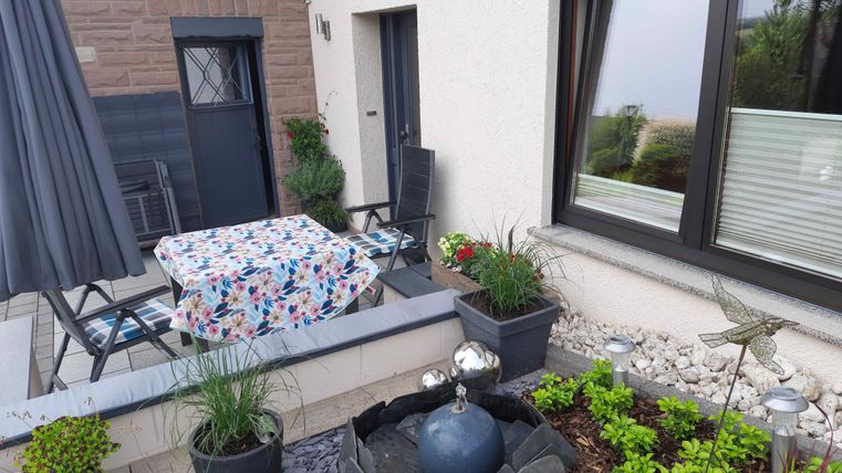 Eine gemütliche Terrasse mit einem Tisch und Stühlen, bedeckt mit einem bunten Tischdecken. Umgeben von blühenden Pflanzen und dekorativen Elementen.