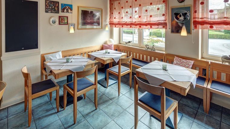 Un restaurant confortable avec des chaises et des tables en bois. De grandes fenêtres avec des rideaux rouges laissent entrer beaucoup de lumière.