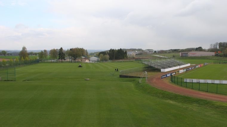 Een groot groen sportveld met gras en een aangrenzend stadion. Op de achtergrond zijn enkele bomen en gebouwen te zien.