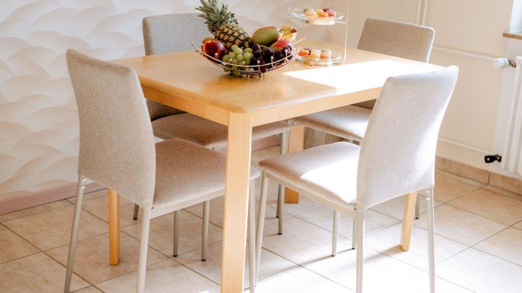 Ein heller Essbereich mit einem Holz-Tisch und vier stuhlähnlichen Sesseln. Auf dem Tisch steht ein Obstkorb mit verschiedenen Früchten.
