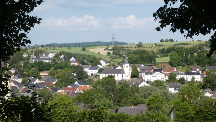 Une vue pittoresque d'un petit village avec des maisons traditionnelles et une église. Les environs sont verts avec des collines douces et un ciel dégagé.
