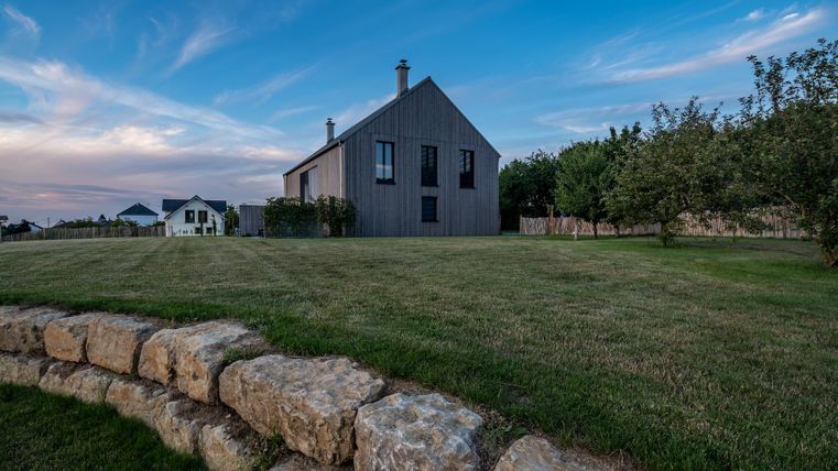 Ein modernes Haus mit einer Holzfassade steht in einer grünen Wiese. Der Himmel ist strahlend blau mit sanften Wolken.