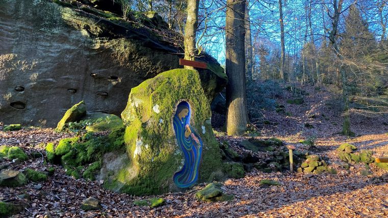 Ein bunter Fels mit einer künstlerischen Darstellung in einem Wald. Umgeben von Laub und hohen Bäumen im Sonnenlicht.