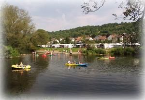 Un paysage aquatique calme avec plusieurs canoës. À l'arrière-plan, on peut voir quelques maisons et arbres.