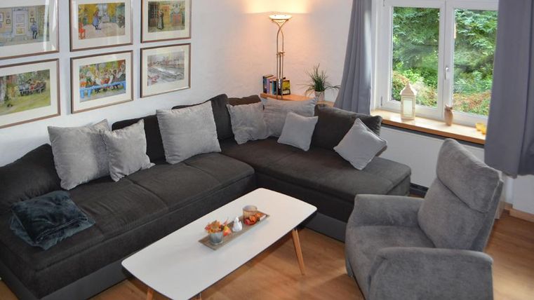 Ein gemütliches Wohnzimmer mit einem großen, grauen Sofa und einem Sessel. Neben einem Fenster stehen ein Tisch und einige dekorative Kissen, alles wirkt einladend.