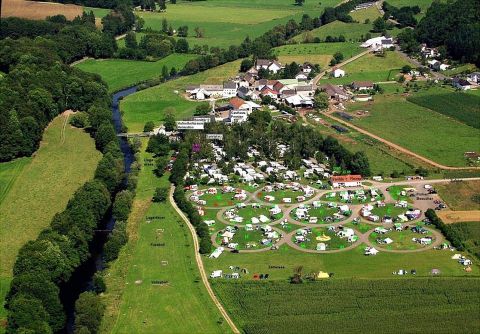 Een luchtfoto van een camping met veel tenten en campers in een groene landschap. Op de achtergrond stroomt een kleine rivier en er zijn enkele gebouwen te zien.