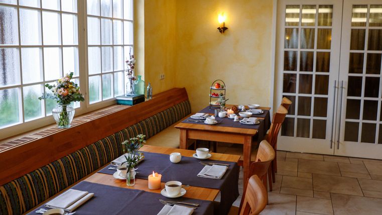 Un coin repas confortable avec des tables et chaises en bois. Les fenêtres laissent entrer beaucoup de lumière, et des fleurs et des bougies sont disposées sur les tables.