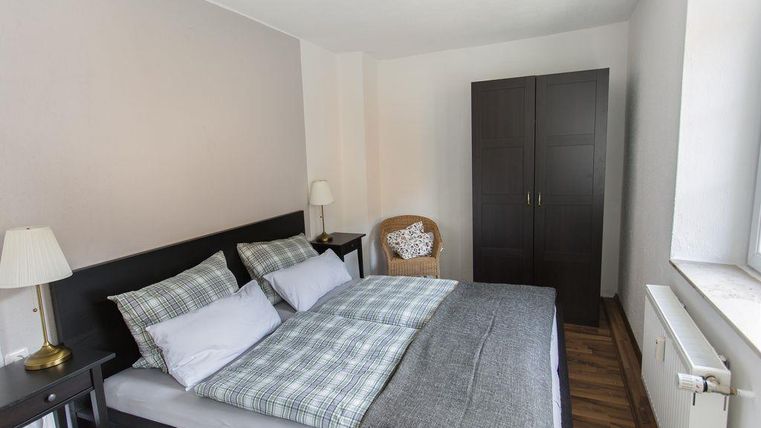 A cozy bedroom with a large bed and pillows in gray and checked. There is a chair, a lamp, and a wardrobe.