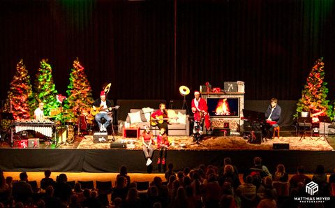 Ein musikalisches Weihnachtskonzert auf der Bühne mit mehreren Musikern und Weihnachtsdekoration. Im Hintergrund sind festlich geschmückte Tannenbäume zu sehen.