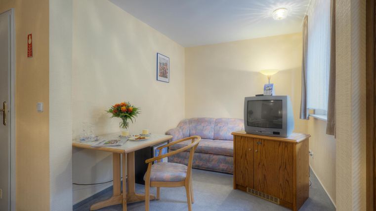 Ein gemütlicher Wohnbereich mit einem Tisch, einem Stuhl und einem Sofa. Ein Fernseher steht auf einem kleinen Schrank und frische Blumen sind auf dem Tisch.