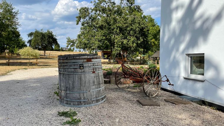 Ein Holzfass steht neben einem alten Wagen in einem ländlichen Garten. Im Hintergrund sind Bäume und ein helles Gebäude zu sehen.