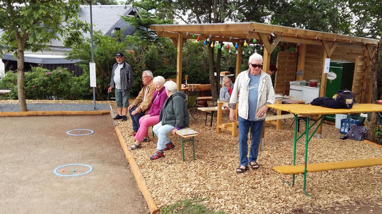 Eine Gruppe älterer Menschen sitzt und steht in einem Gartenbereich. Im Hintergrund befindet sich eine überdachte Sitzgelegenheit und ein Spielplatz.